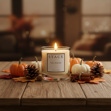 Load image into Gallery viewer, A lit candle in a clear glass jar sits on a rustic wooden table surrounded by small pumpkins, pinecones, cinnamon sticks, and autumn leaves. The candle label reads 'STACK FALL HARVEST' and 'LONDON CANDLE.