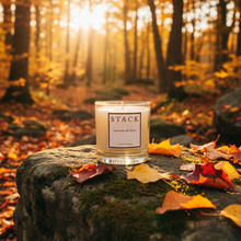 Load image into Gallery viewer, A glass jar candle labeled STACK Candle of Time sits on a mossy stone surrounded by fallen autumn leaves in a sunlit forest.